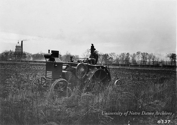 A worker on a tractor plows a Notre Dame farm field in the spring of 1924.  The Main Building and Power Plant smoke stack are in the background.