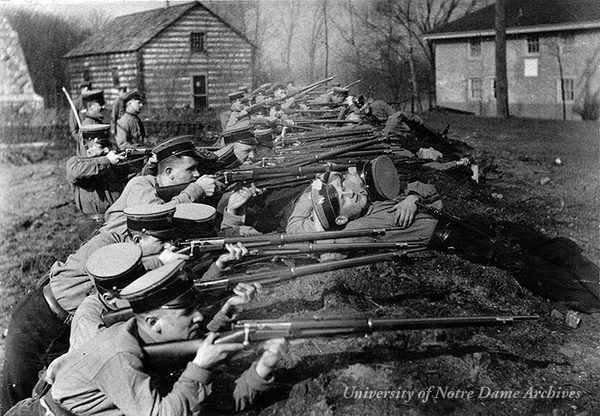 Students in the military training corps at rifle target practice in the trenches, 1915-1916.  The Founder's Monument, Log Chapel, and Old College are in the background.