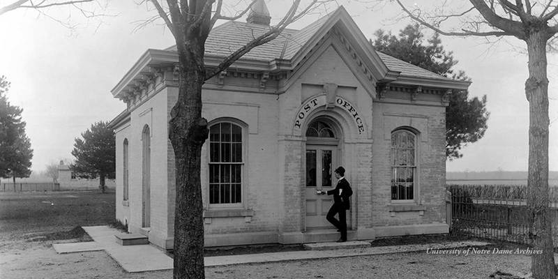A man dropping a letter into the mail slot of the front door of Notre Dame's first Post Office built in 1856, located east of the present location of the Sorin Statue on the Main Quad, c1890s.